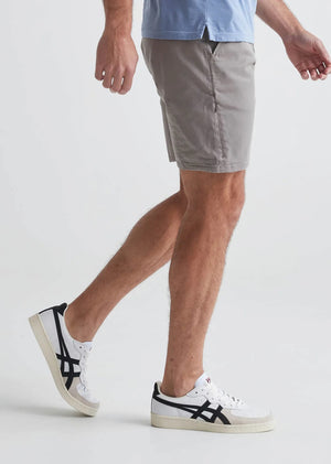 A person faces away from the camera, wearing a white T-shirt, DUER Live Free Journey Shorts in Titanium (slim fit), and white sneakers with black heel tabs, set against a plain white background.