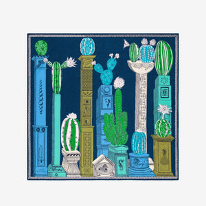 A woman with dark hair tied back wears a white outfit, accessorized with the INOUI EDITIONS - TEQUILA SCARF IN CURACAO by INOUI, featuring green, yellow, and blue patterns, set against a plain white background.