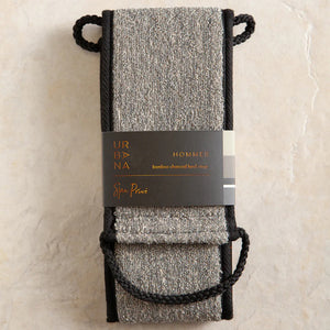 The BAMBOO CHARCOAL BACK SCRUBBER by EUROPEAN SOAPS, LLC is a rectangular, grey synthetic cleaning cloth with black trim. It is folded over and features black looped handles on both ends for an easy grip during use. The texture of the cloth appears to be suitable for gentle cleaning or polishing dead skin cells away.