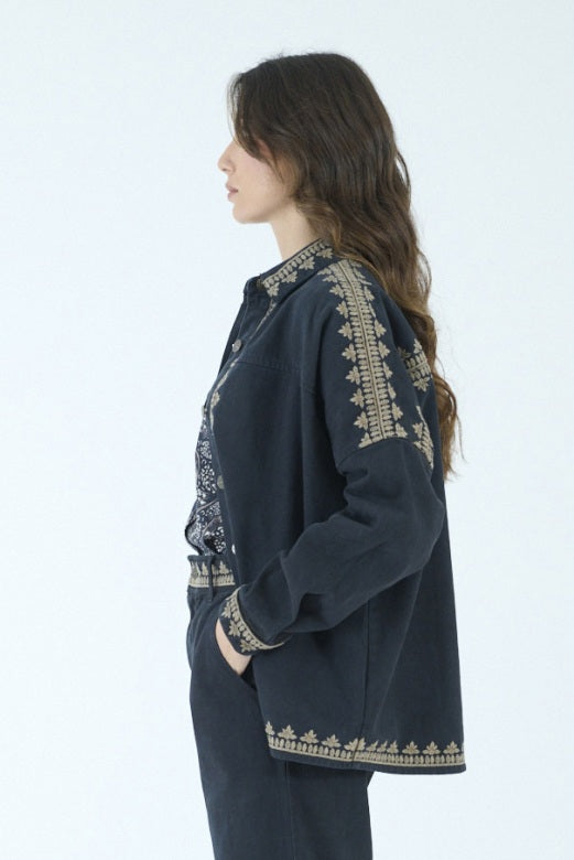 A woman with long brown hair stands indoors in front of large windows, embodying effortless style in the STORIATIPIC ROXY JACKET IN NOIR by STORIATIPIC, featuring gold embroidery on 100% cotton. She pairs the jacket with a patterned shirt, dark pants, and black loafers, mindful to follow proper cotton care such as gentle machine wash.