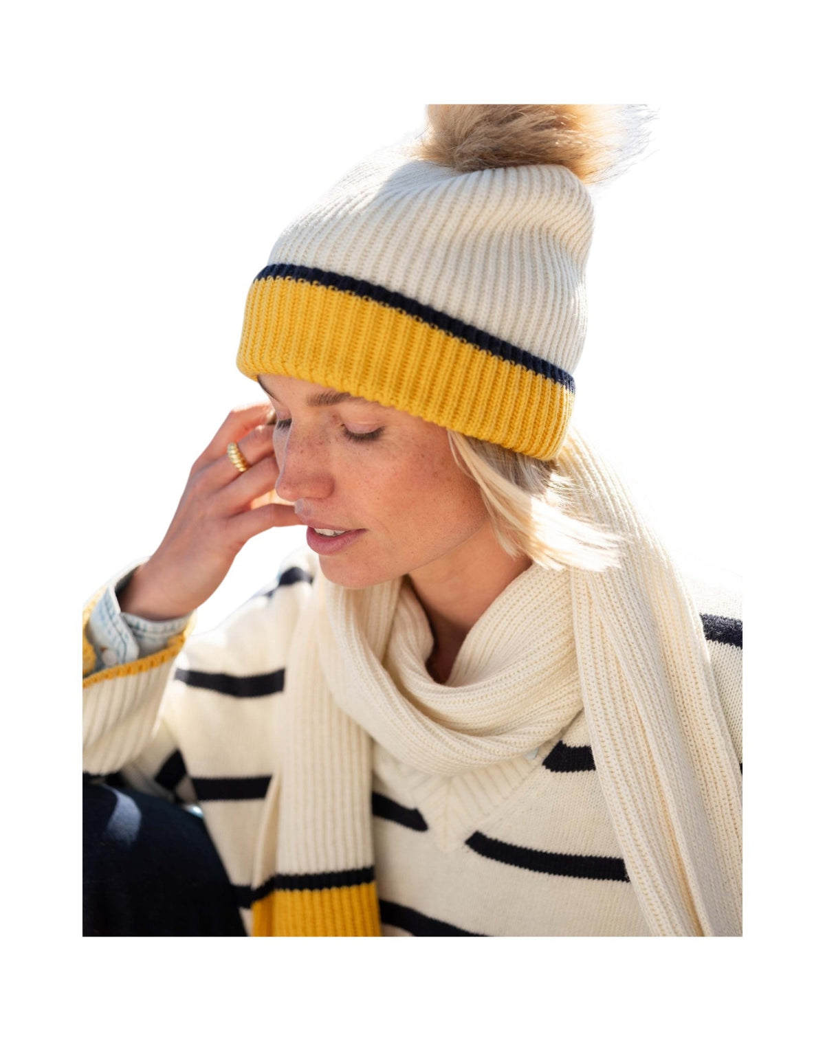 A woman, framed against a white background, wears the MERSEA - MONTAUK COLORBLOCK BEANIE IN CREME STRIPE—a cream knit hat with a yellow stripe and pom-pom—paired with a matching cream scarf and a chunky rib knit sweater accented by black stripes, showcasing the ultimate cold-weather accessory ensemble from MERSEA.