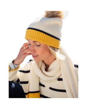 A woman, framed against a white background, wears the MERSEA - MONTAUK COLORBLOCK BEANIE IN CREME STRIPE—a cream knit hat with a yellow stripe and pom-pom—paired with a matching cream scarf and a chunky rib knit sweater accented by black stripes, showcasing the ultimate cold-weather accessory ensemble from MERSEA.