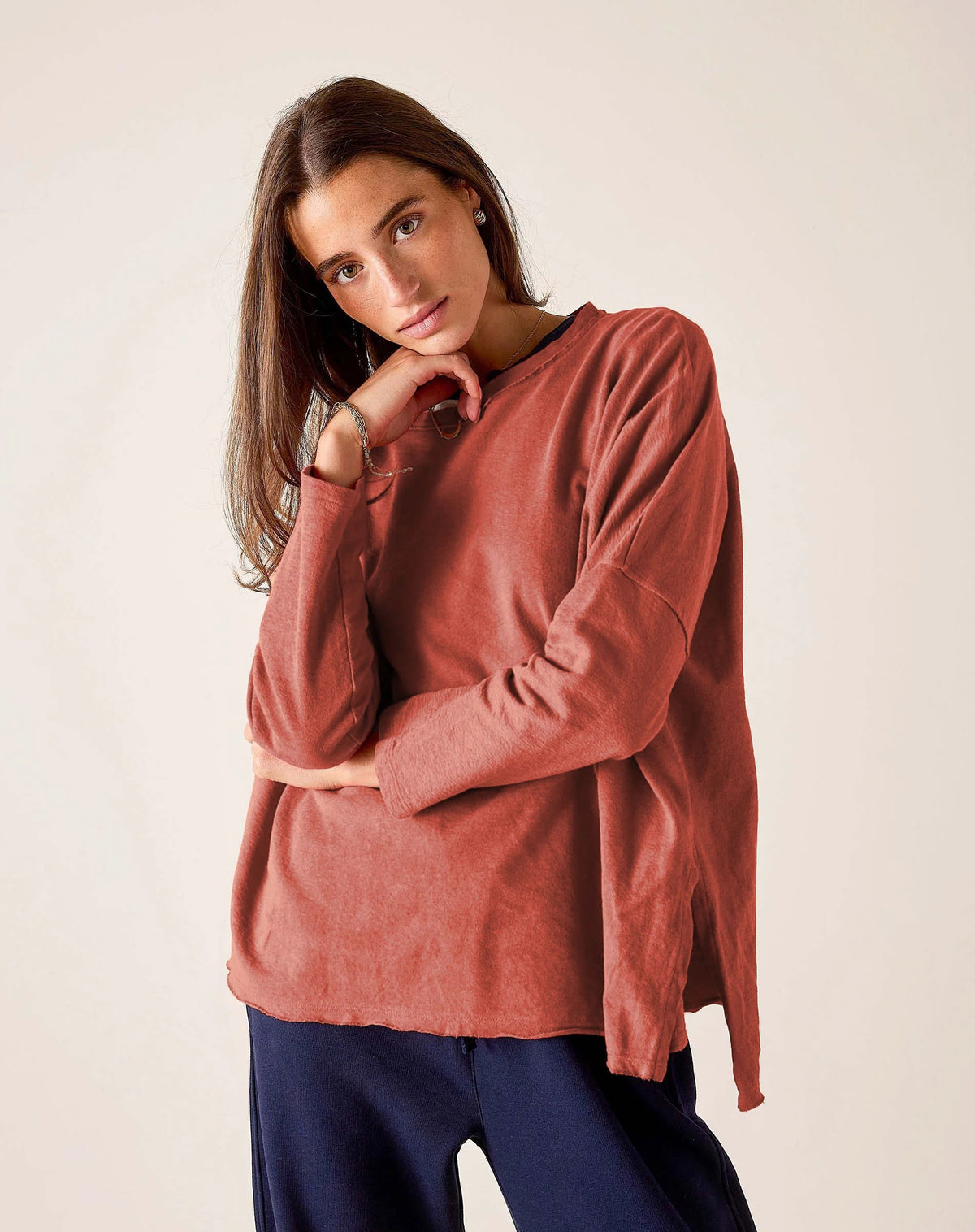 A woman with long brown hair wears the MERSEA Catalina Rustic Tee in Cayenne, paired with dark blue pants. She stands against a plain light background, resting her chin on her hand and gazing at the camera.