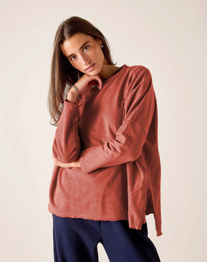 A woman with long brown hair wears the MERSEA Catalina Rustic Tee in Cayenne, paired with dark blue pants. She stands against a plain light background, resting her chin on her hand and gazing at the camera.