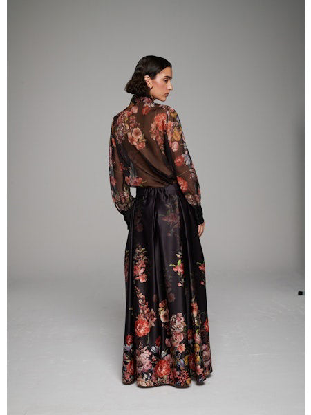 A woman poses in the MILVA MI - LONG FLORAL MAXI SKIRT by MILVA, paired with a sheer, long-sleeved floral blouse and pointed black heels against a simple studio backdrop.