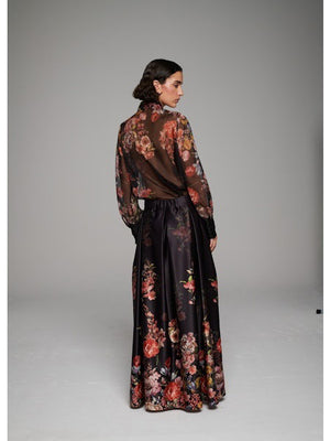 A woman poses in the MILVA MI - LONG FLORAL MAXI SKIRT by MILVA, paired with a sheer, long-sleeved floral blouse and pointed black heels against a simple studio backdrop.