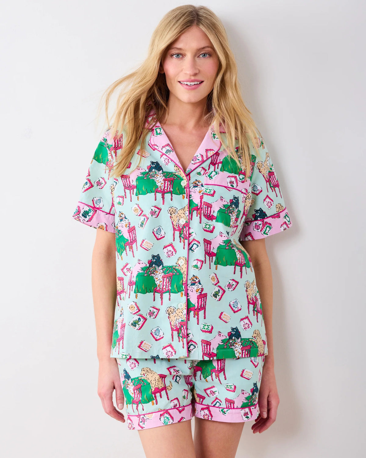 Dressed in PRINTFRESH’s Mahjong Menagerie Pajama Short Set, this person sports matching pastel organic cotton sleepwear featuring a playful cat print with tables, books, and chairs. The set includes a short-sleeve button-up shirt and shorts, both finished with light pink trim.