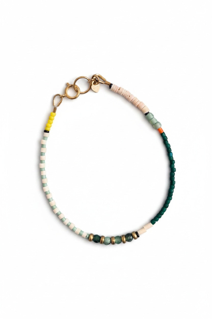 The ALICE RISE - EMERALD BRACELET by ALICE RISE showcases handmade North Carolina artistry with white, yellow, green, teal, and metallic gold beads and a gold clasp, elegantly displayed on a textured blue surface.