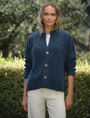 A person with wavy blonde hair, wearing a relaxed fit FRANK & EILEEN Montecito Crew Neck Cardigan in Pure Italian Cotton Blueberry and white pants, stands outdoors facing away, surrounded by greenery and trees.