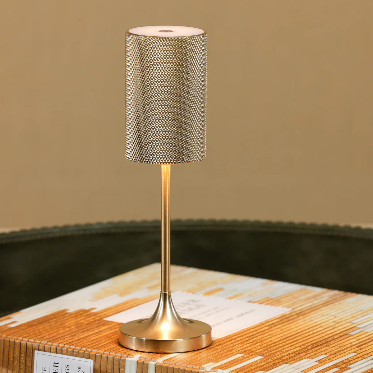 The FACET RECHARGEABLE LAMP by ZODAX is a modern gold table lamp with a textured cylindrical shade, slender stem, and round base. It offers adjustable light intensity and is displayed against a plain white background.