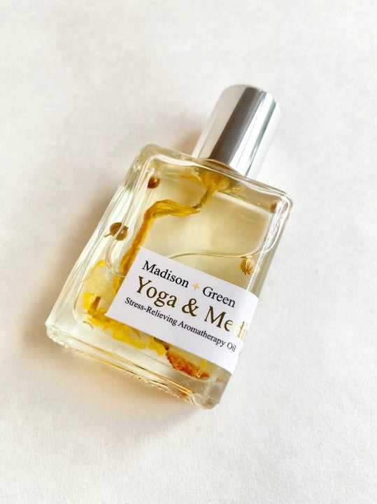 A clear glass bottle with a silver cap holds yellow aromatherapy oil labeled “MADISON AND GREEN - YOGA & MEDITATION SPIRITUAL AROMATHERAPY BODY OIL MINI” from MADISON GREEN, with visible herbs or botanicals suspended inside—making this botanical body oil ideal for self-care rituals.
