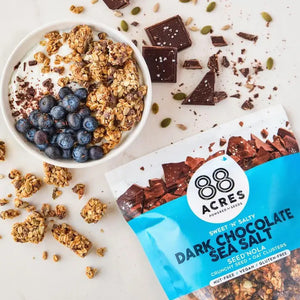 A bag of 88 ACRES - DARK CHOCOLATE SEA SALT SEED'NOLA sits on a light surface. Its blue and white packaging highlights vegan, nut-free seeds, gluten-free oats, and dark chocolate with granola imagery.