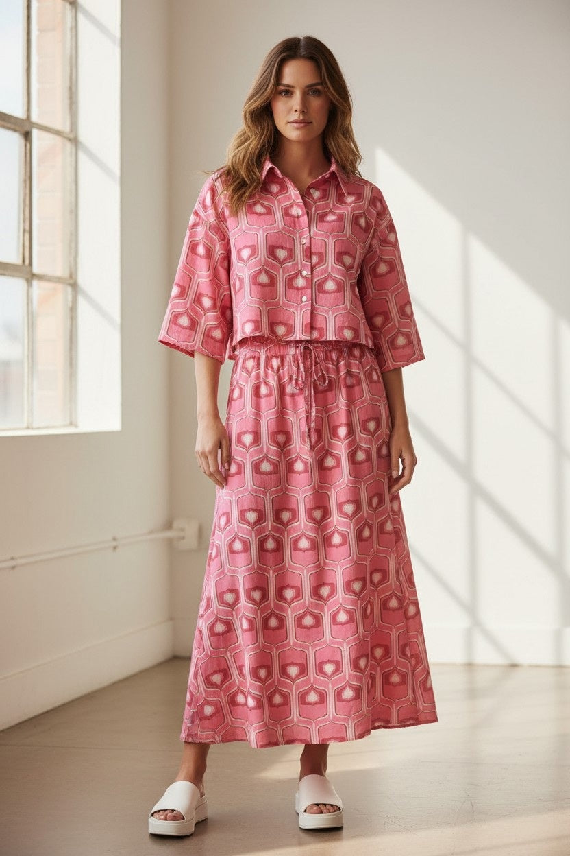 A woman stands indoors by a window, wearing a pink geometric print button-up top paired with the FLOOR - MAXI COTTON SKIRT from FLOOR and white slide sandals. Sunlight streams through the window, illuminating the minimalist room.