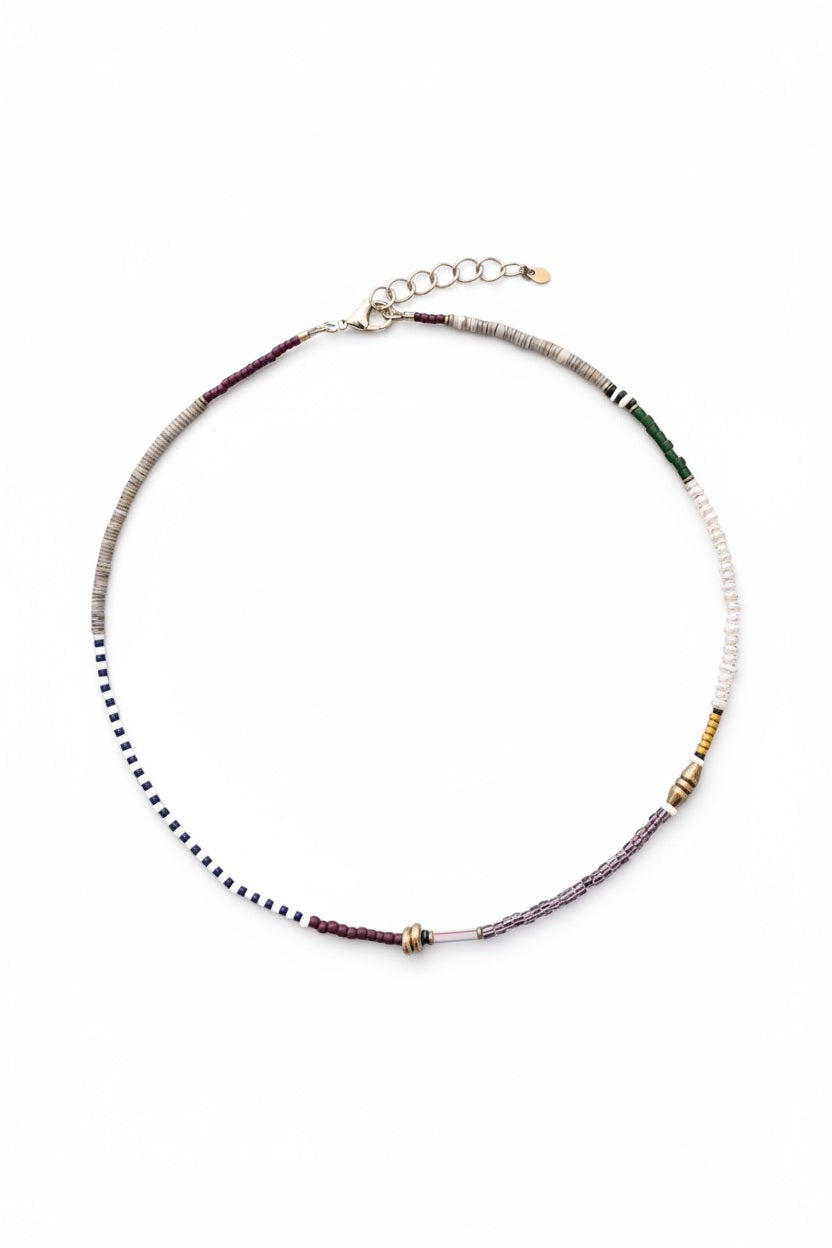 The ALICE RISE - PEARL MIXED MEDIA NECKLACE by ALICE RISE features small, multicolored vintage glass beads arranged in sections and finished with a 14k gold-filled clasp, displayed in a circle on a textured light blue surface.