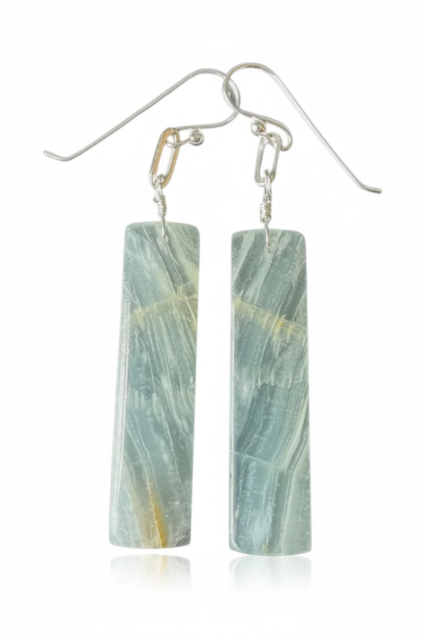 A pair of rectangular blue-green ANN LIGHTFOOT - BLUE CALCITE SLAB EARRINGS, featuring subtle white and yellowish streaks, dangle from sterling silver earwires against a white background.