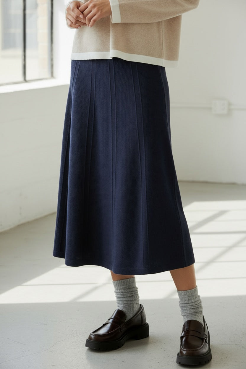 A person, shown from the waist down, wears the ANNECLAIRE knit wool flare skirt in beige with gray socks and black loafers, standing against a light plain background.