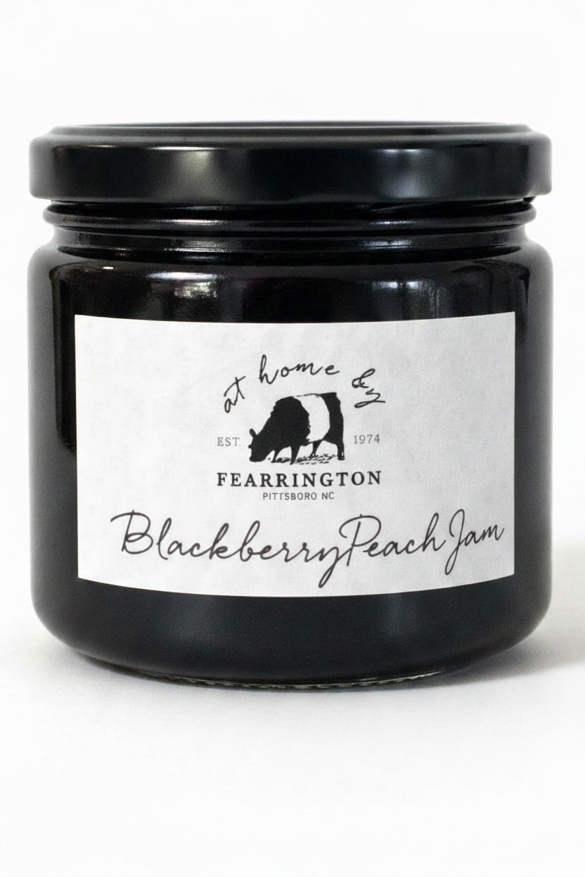 A glass jar of FEARRINGTON SIGNATURE COLLECTION - FEARRINGTON HOUSEMADE BLACKBERRY PEACH JAM with a black lid and a white label. The label lists ingredients: blackberries, peaches, lemon, cinnamon, nutmeg, sugar, pectin. It also mentions: "Handmade with care in our kitchens by Fearrington chefs." Perfect as a hostess gift or for breakfast jams. May contain allergens.