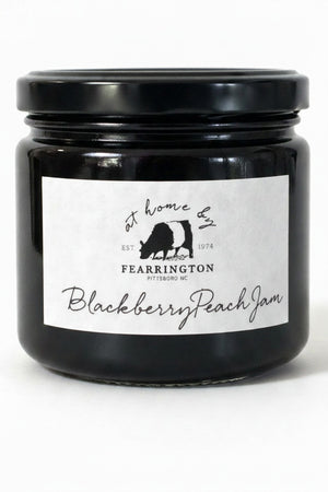 A glass jar of FEARRINGTON SIGNATURE COLLECTION - FEARRINGTON HOUSEMADE BLACKBERRY PEACH JAM with a black lid and a white label. The label lists ingredients: blackberries, peaches, lemon, cinnamon, nutmeg, sugar, pectin. It also mentions: "Handmade with care in our kitchens by Fearrington chefs." Perfect as a hostess gift or for breakfast jams. May contain allergens.