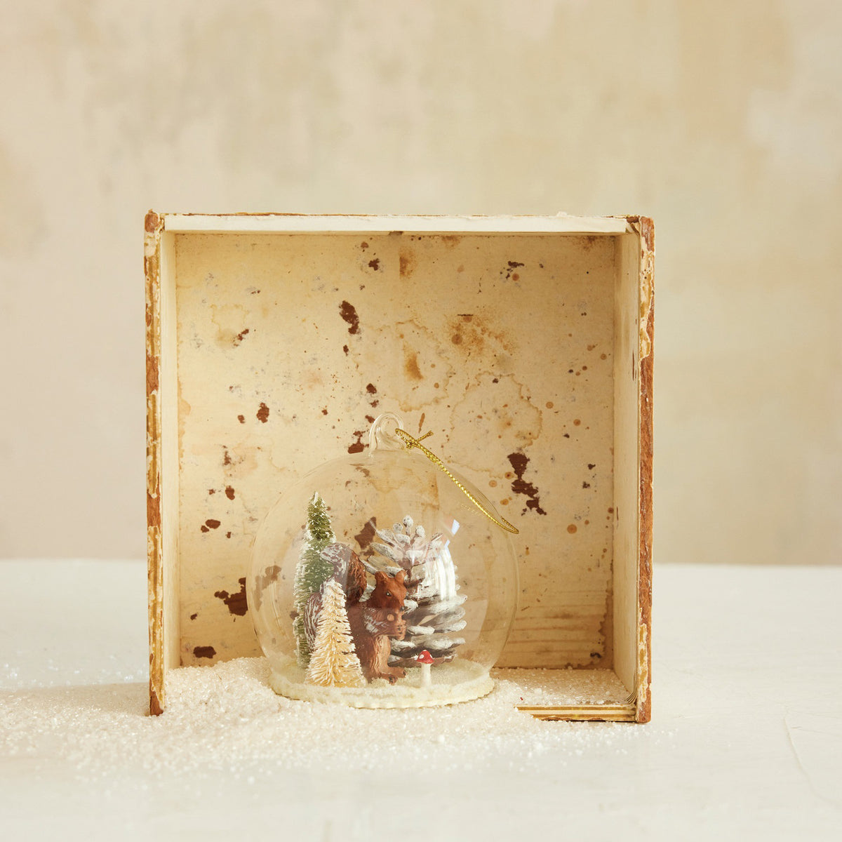 The CREATIVE COOP SNOW GLOBE ORNAMENT WITH WOODLAND SQUIRREL is a clear glass ornament with a gold hanging string, showcasing a pinecone, squirrel, and snowy ground scene inside.