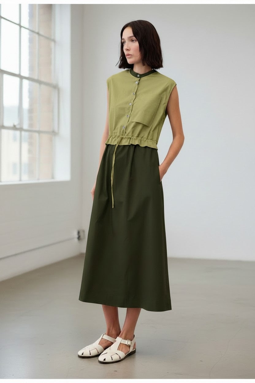 A woman stands in a minimal room wearing the FLOOR Colorblock Cotton Dress in Green by FLOOR. The sleeveless dress features olive green cotton poplin with buttons, and she pairs it with white sandals. Her dark shoulder-length hair frames her face as she keeps her hands in her pockets.