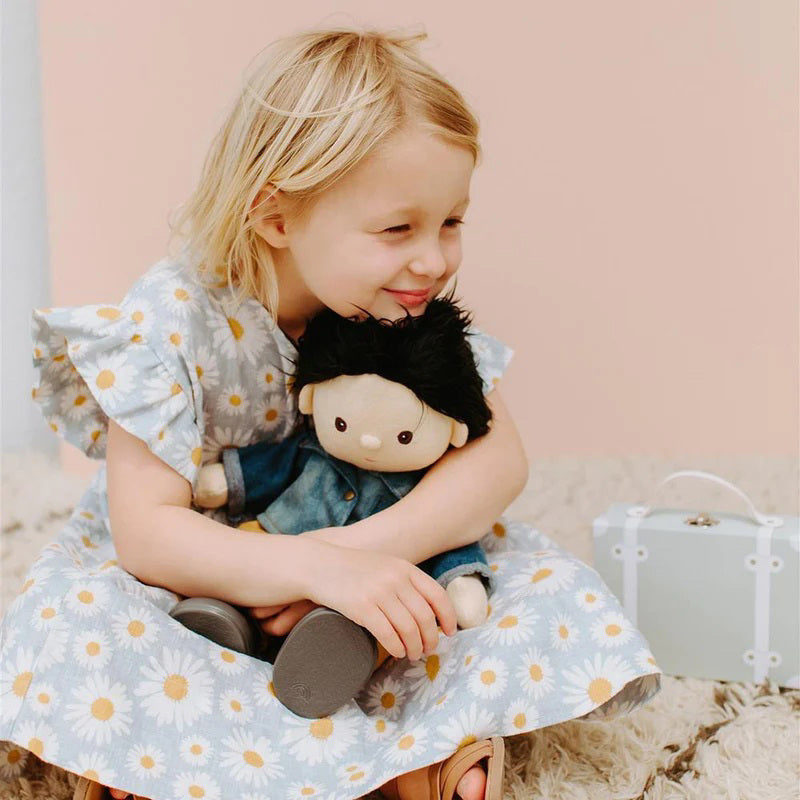 A Dinkum Doll from Olli Ella USA's Travel Togs collection, featuring dark hair and dressed in a denim jacket, mustard pants, and black shoes, stands on a wooden surface. Beside the doll is a teal See-Ya Suitcase with white trim and hardware. In the background, another larger teal suitcase is partially visible.