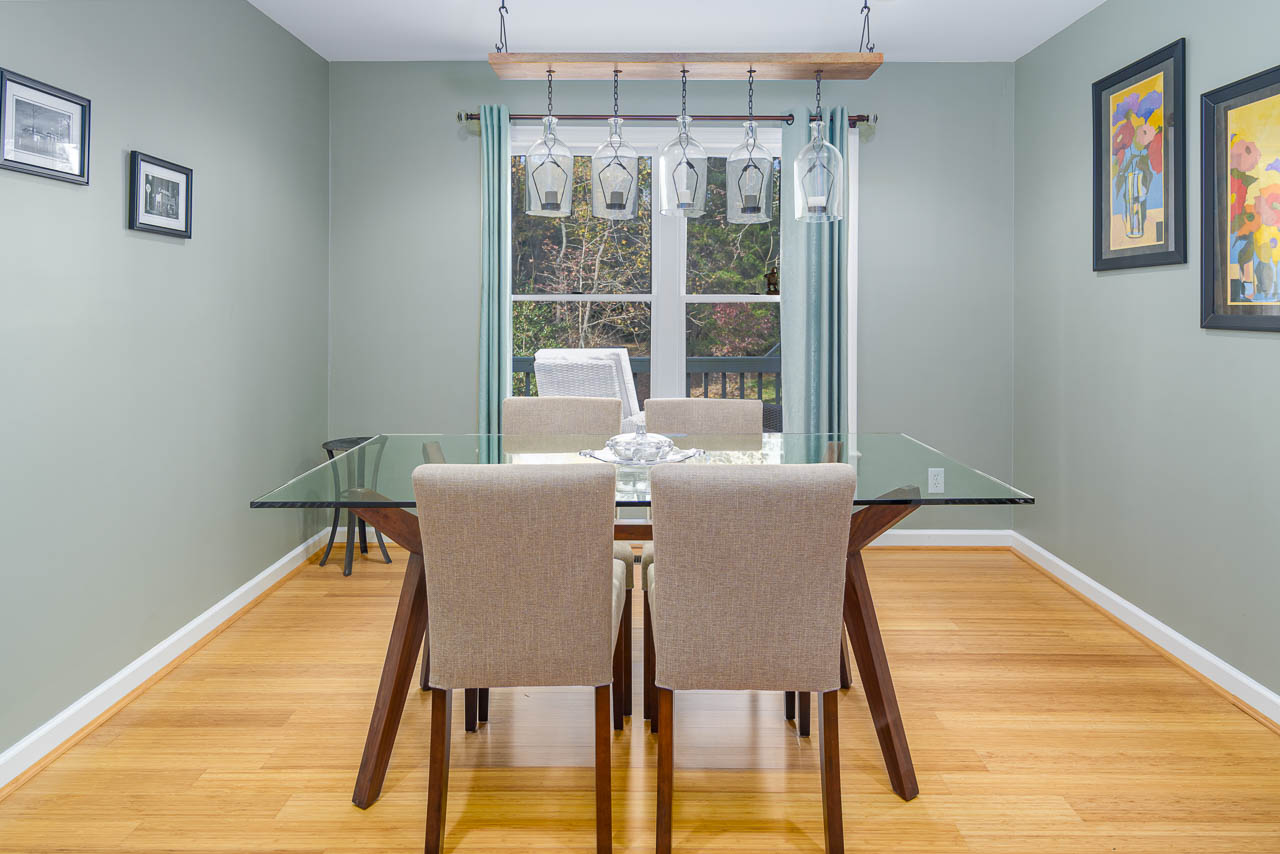 A modern dining room with a glass table, four beige chairs, and a contemporary rectangular pendant light. The room has light wood floors, green walls, and is decorated with framed art and a large window with green curtains.