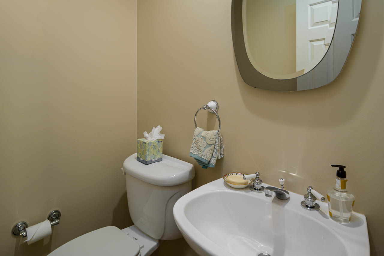 A small bathroom with beige walls features a white toilet, a box of tissues, a towel ring with a decorative towel, a white sink with a mirror above, soap dispenser, and a roll of toilet paper on a holder.