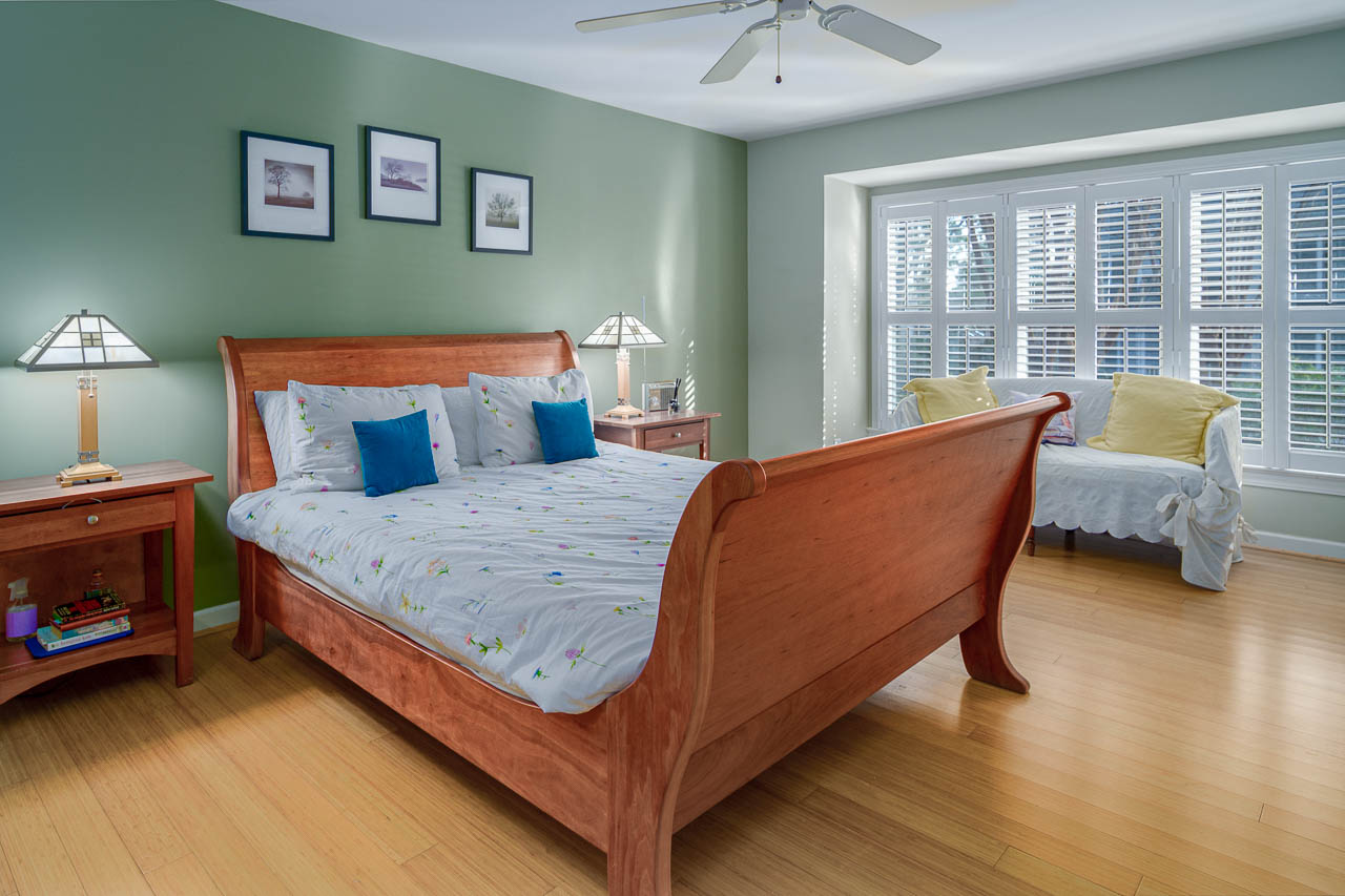 Bedroom with a wooden sleigh bed, two bedside tables with lamps, framed pictures on a green wall, wooden floor, a window with white shutters, and a small bench with cushions.