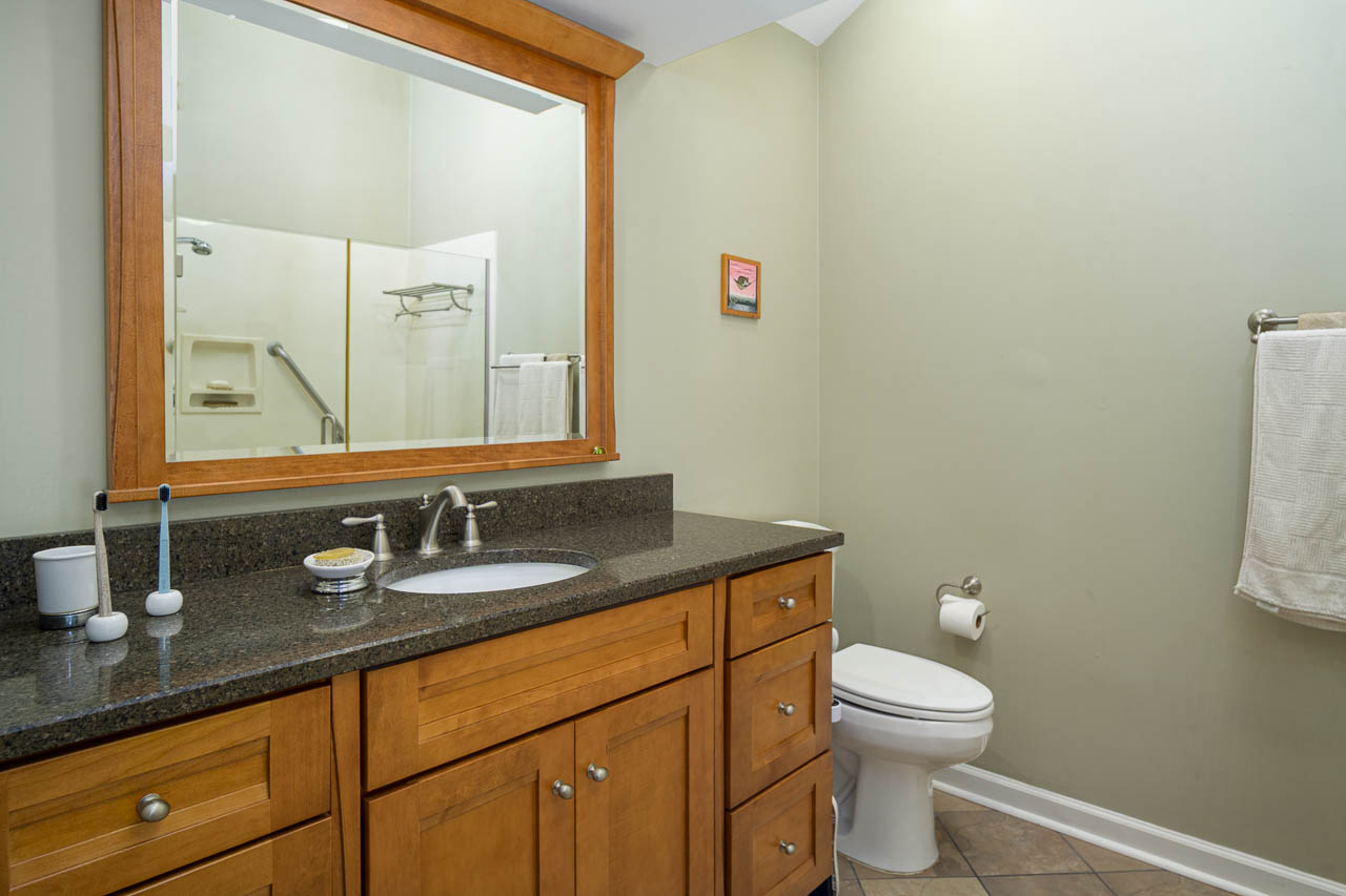 A modern bathroom with light wood cabinets, a black countertop, a sink with a silver faucet, a large mirror, a toilet, a towel rack with a white towel, and a shower visible in the background.