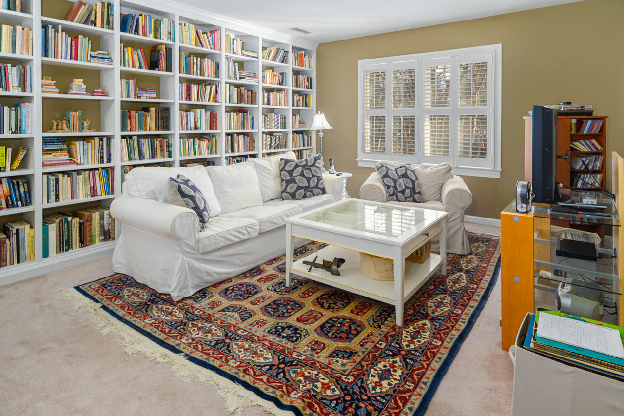 A cozy living room with white sofas, a glass coffee table, and a patterned rug. Floor-to-ceiling bookshelves filled with books line one wall. A window with shutters lets in natural light. A TV and shelves are on the right.