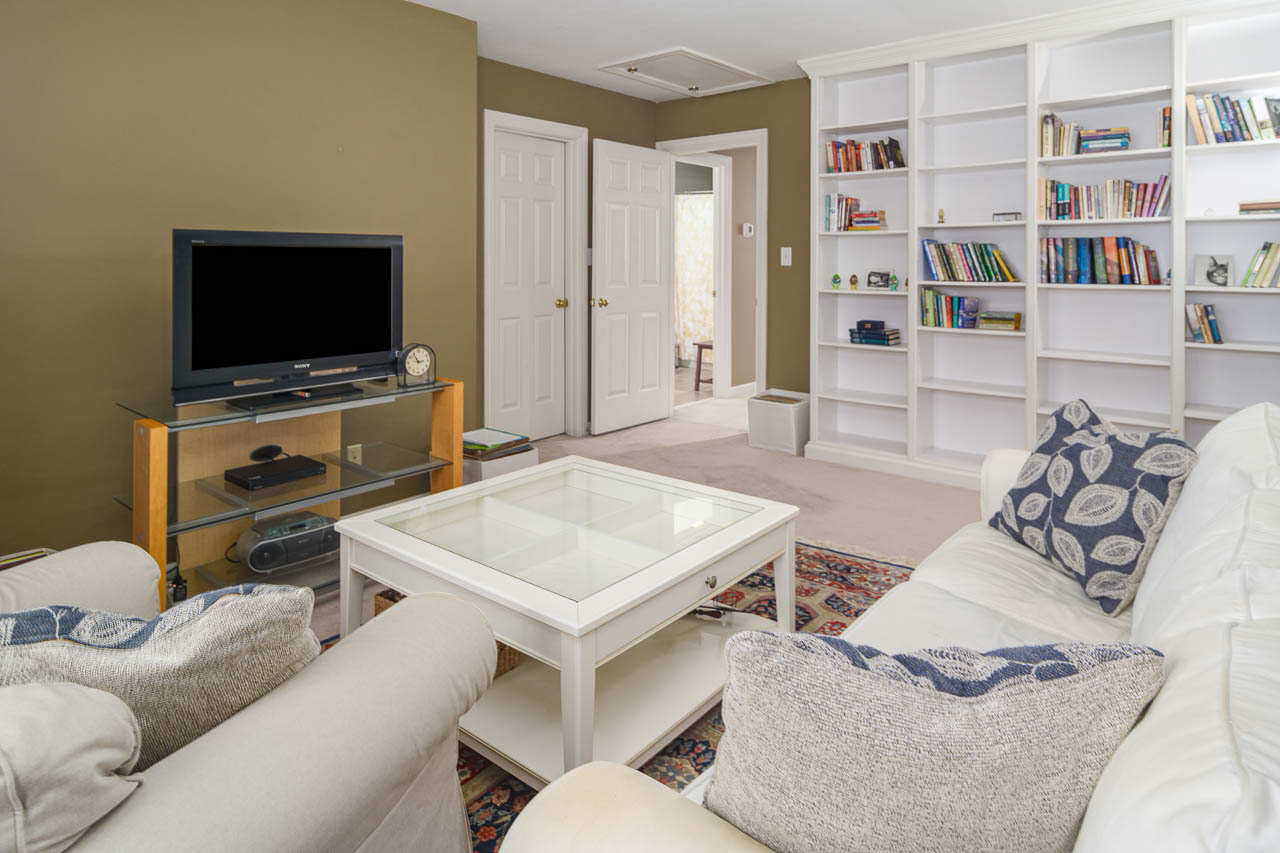 A cozy living room with a white sofa, patterned cushions, a glass coffee table, a TV on a stand, and built-in white bookshelves filled with books against a beige wall. Two closed white doors are visible in the background.