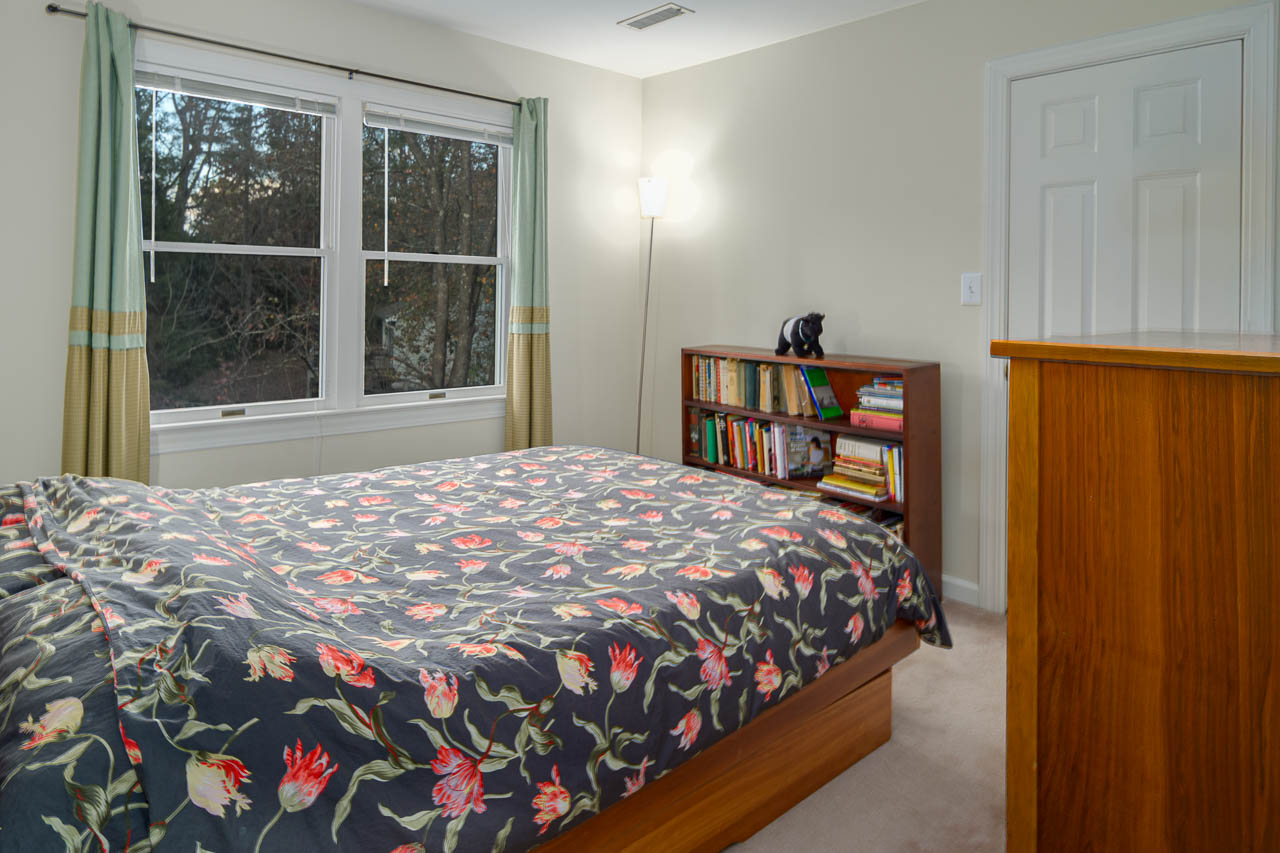 A cozy bedroom with a floral-patterned bedspread, wooden dresser, bookshelf filled with books, large window with green curtains, and a floor lamp in the corner.