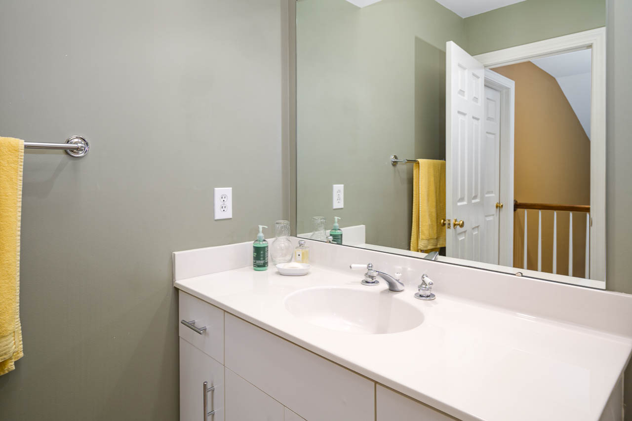 A bathroom with a white sink and countertop, a large mirror, soap dispensers, and yellow towels. The walls are light green, and an open door reveals a hallway with tan walls and a wooden railing.