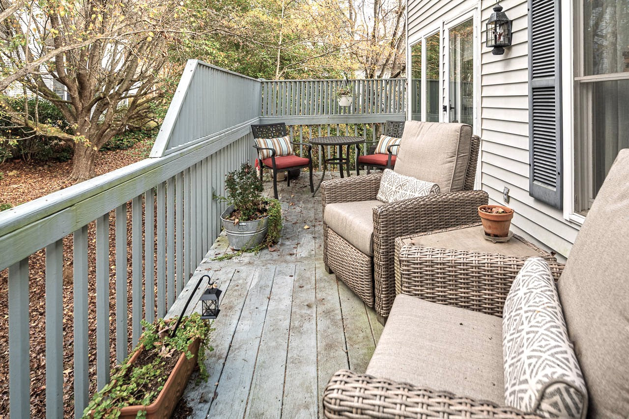 A cozy wooden deck with wicker chairs, a small table with two chairs, potted plants, and railings, next to a house with light siding. Trees and leaves are visible in the background.