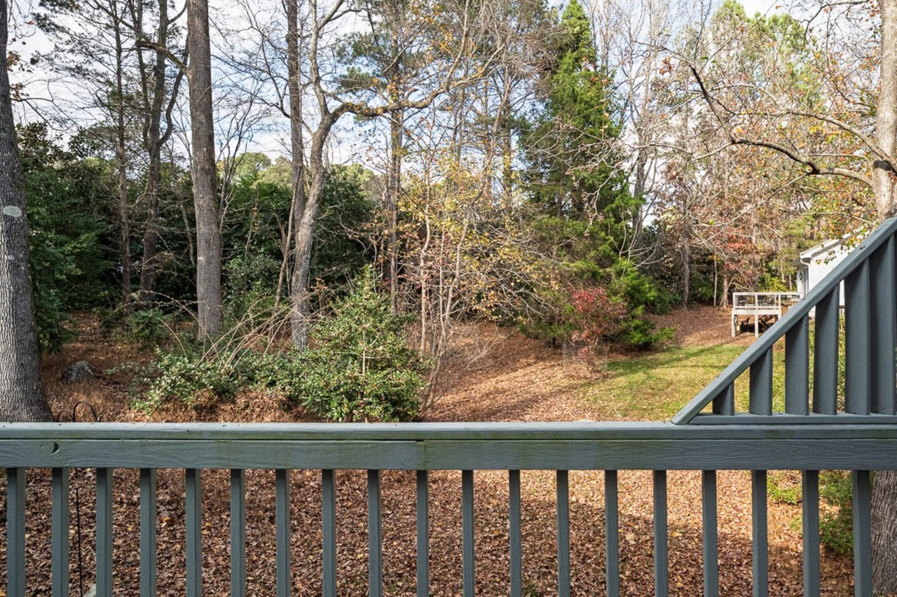 View from a deck with a wooden railing overlooking a wooded backyard with trees, fallen leaves, and a small patch of green grass; a shed is visible in the distance.