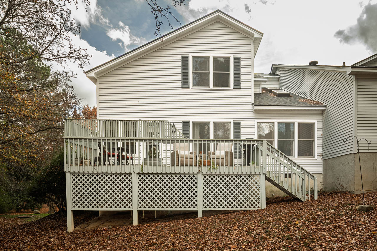 A white two-story house with a wooden deck and stairs leading to the yard, surrounded by autumn trees and scattered brown leaves on the ground.