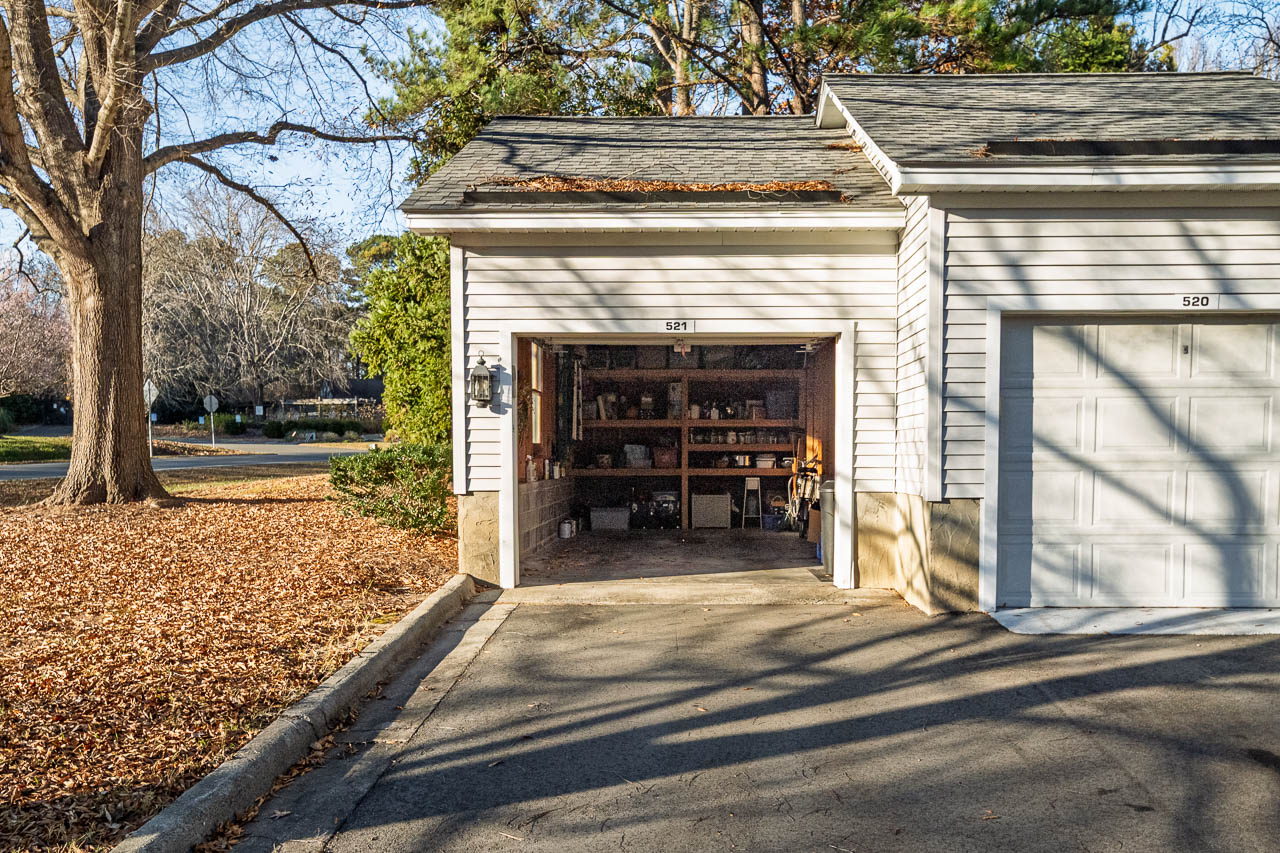 A single-car garage with the door open reveals shelves with tools and supplies. Fallen autumn leaves cover the driveway and yard. Sunlight casts long shadows from nearby trees.