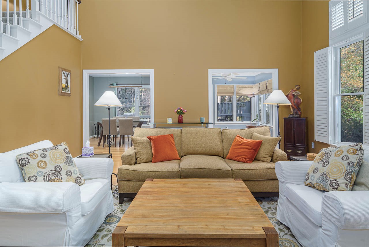 A cozy living room with a tan sofa, two white armchairs, and a wooden coffee table. The walls are mustard yellow, and there are orange pillows and patterned cushions. Large windows and an open view to the dining area are visible.