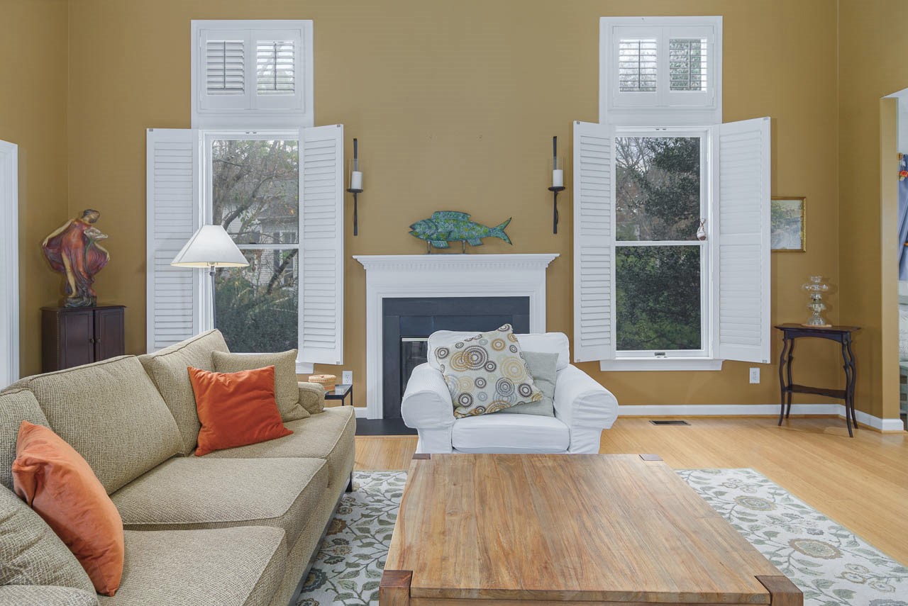 A cozy living room with a beige sofa and orange pillows, a white armchair with a patterned pillow, a wooden coffee table, a fireplace with white shutters, and decorative art pieces on the mantel and side table.