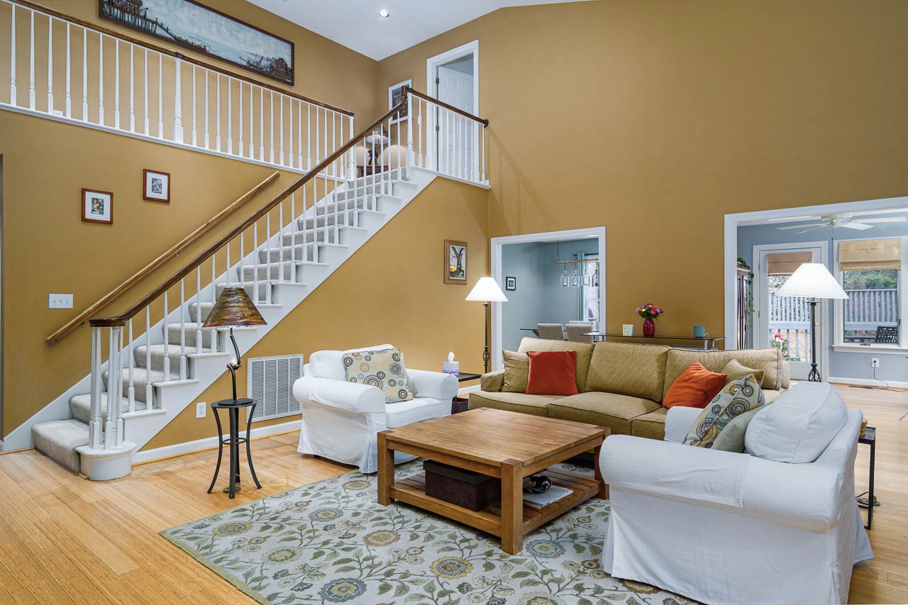 A cozy living room with mustard yellow walls, a beige sectional sofa, two white armchairs, a wooden coffee table, and a patterned rug. A staircase with white railings leads to an upper level. The room is well-lit and inviting.
