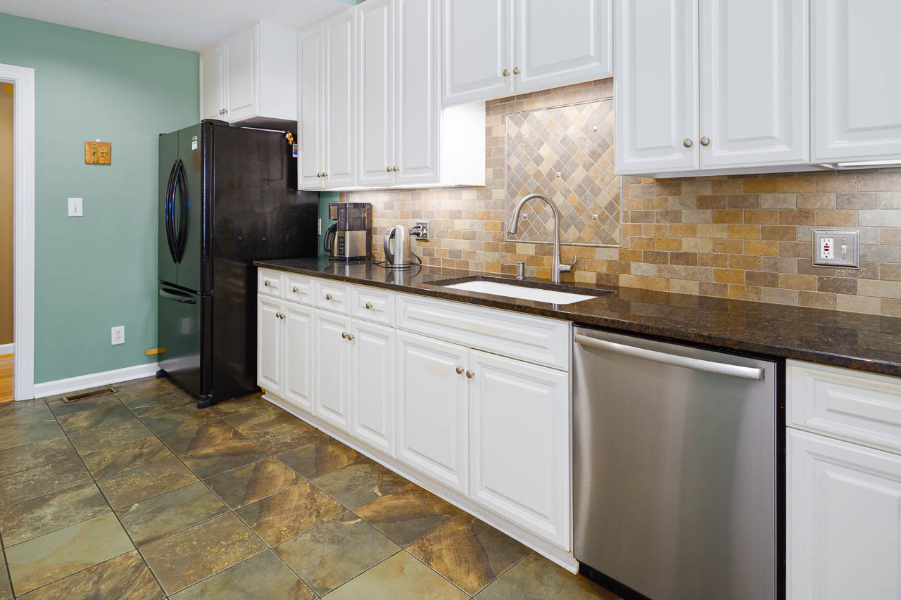 A modern kitchen with white cabinets, a black refrigerator, stainless steel dishwasher, brown stone tile backsplash, and greenish-brown tiled floor. The countertop is dark and the walls are painted light green.