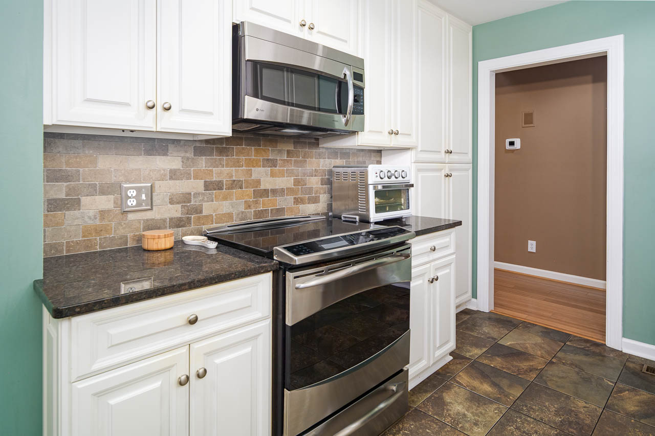 Modern kitchen with white cabinets, stainless steel oven, microwave, and toaster oven. Brown tile backsplash, dark granite countertop, and tiled floor. Green painted walls and an open doorway to a hallway.