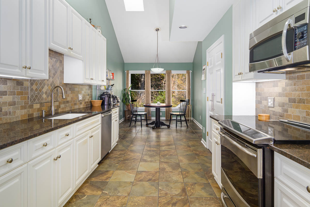 Modern kitchen with white cabinets, stainless steel appliances, tile backsplash, and brown granite countertops; a round dining table with chairs is set by large windows at the far end of the room.