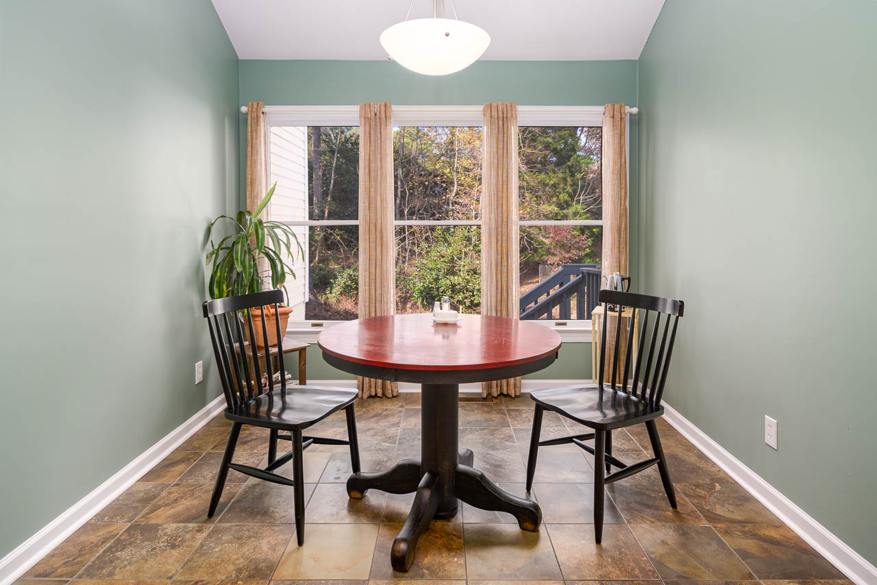 A small dining area with a round wooden table, two black chairs, tile flooring, green walls, a potted plant in the corner, and large windows with beige curtains overlooking a wooded backyard.