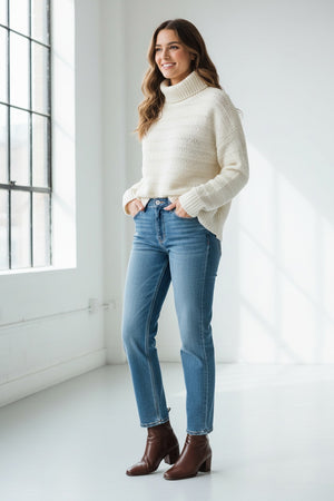A person with curly hair wears the & ISLA Textured Mock Neck Pullover in Ivory and blue jeans, standing against a plain white background, looking confidently at the camera.