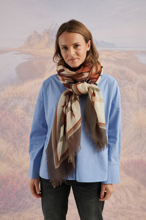 A woman in a blue shirt and jeans stands holding the STORIATIPIC - JULIE WOOL SCARF, a luxury plaid accessory in earthy tones. Behind her, mountains, beach, and grassy dunes create a scenic backdrop.