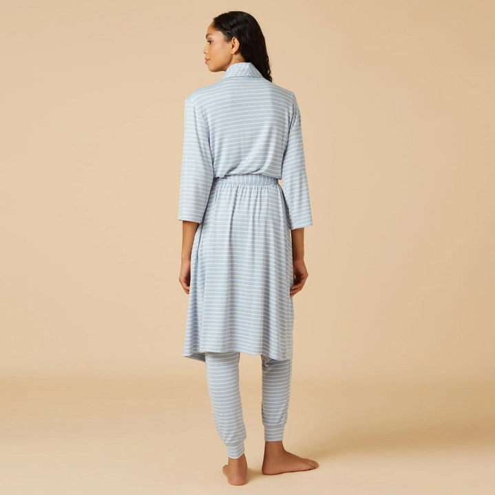 Wearing light blue and white striped pajamas, a person stands barefoot against a beige background, draped in the SOFTIES Dream Shawl Collar Robe in Sky Blue Stripe. This lightweight robe from SOFTIES adds an extra layer of silky smooth comfort to the cozy look.