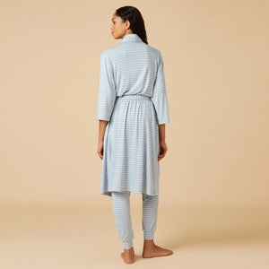 Wearing light blue and white striped pajamas, a person stands barefoot against a beige background, draped in the SOFTIES Dream Shawl Collar Robe in Sky Blue Stripe. This lightweight robe from SOFTIES adds an extra layer of silky smooth comfort to the cozy look.