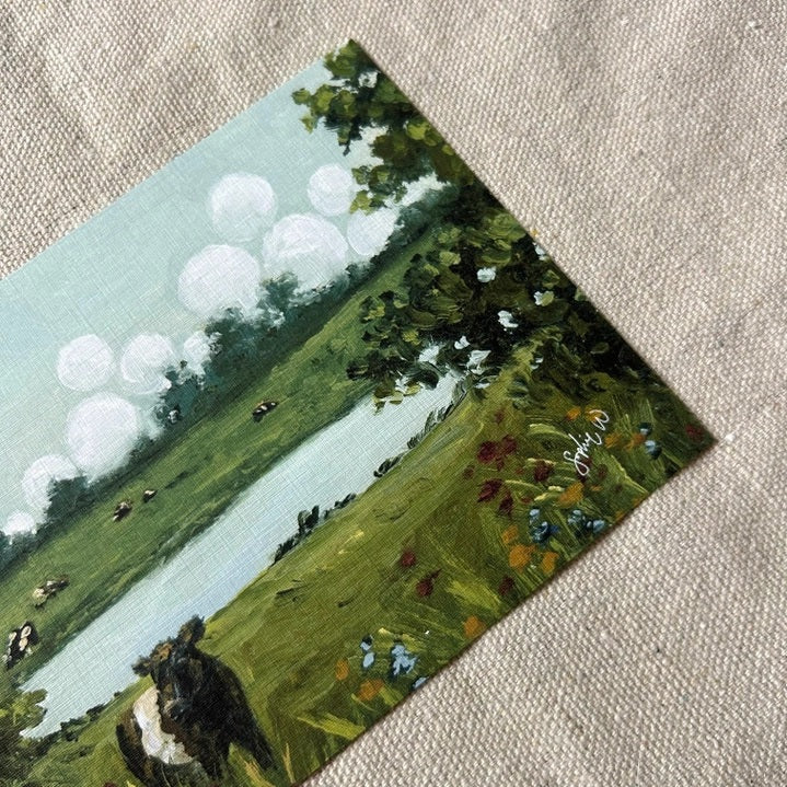 Bring the countryside to your home with the SOPHIE WYATT STUDIO - WILD MEADOWS & BELTIES ART PRINT (12x16). This fine art print by SOPHIE WYATT STUDIO depicts a serene scene where a Belted Galloway cow stands amid wildflowers beside a tranquil pond, while more cows graze in the distant field. Lush bushes frame the landscape and white fluffy clouds float across a vivid blue sky, perfectly capturing the peaceful charm of rural life.