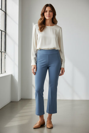 A woman stands indoors by a large window, wearing a long-sleeve cream blouse and the KRAZY LARRY, INC PULL ON WIDE ANKLE PANT in black, which features a tummy tuck design. She completes her look with black loafers and smiles slightly, her wavy brown hair catching the sunlight that casts shadows on the white floor and wall.